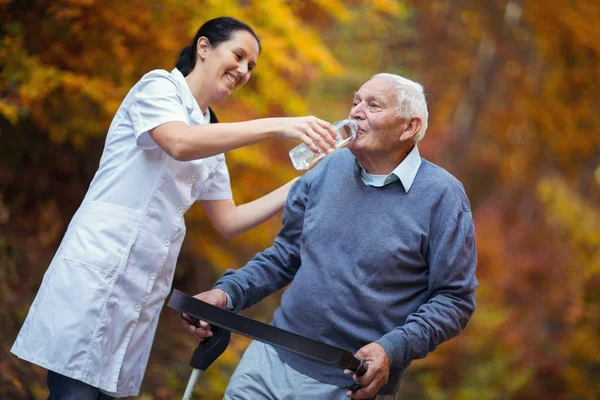 Медицинский работник помогает пожилому человеку с ходячим выпить в парке — стоковое фото