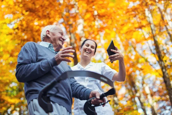 Enfermera cuidadora sonriente y paciente sénior discapacitado en walker usando tableta digital al aire libre —  Fotos de Stock