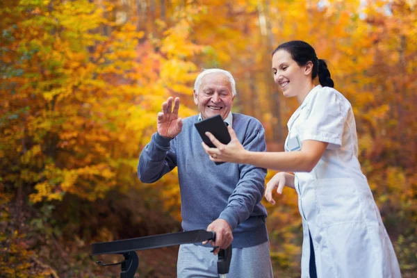 Enfermeira cuidadora sorridente e paciente idoso deficiente em andador usando tablet digital ao ar livre — Fotografia de Stock