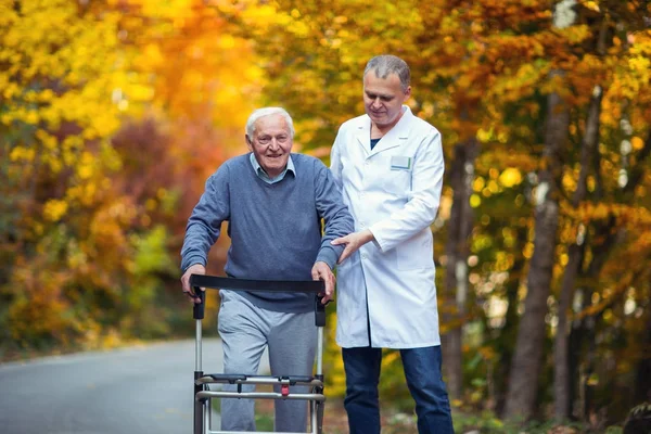 Мужчина-медбрат помогает старшему пациенту с ходунками на открытом воздухе — стоковое фото