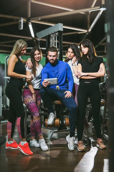 handsome personal trainer using digital tablet, group of sporty women around him in gym