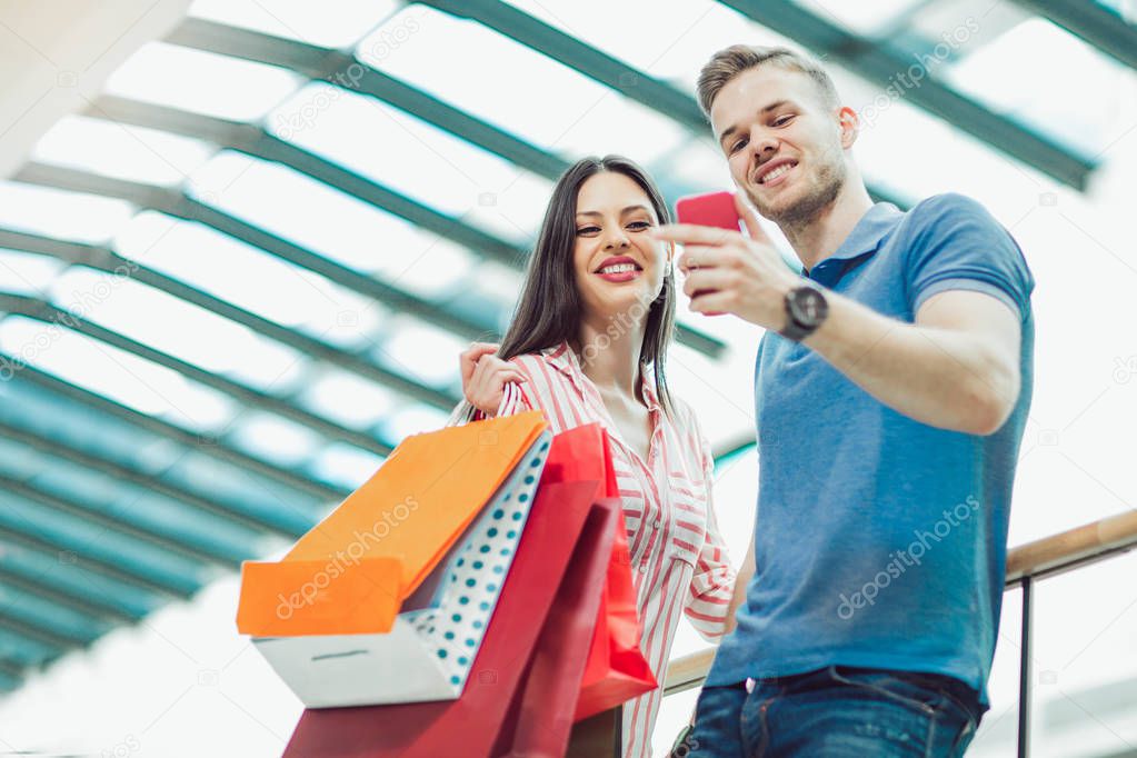 happy young couple with shopping bags and smartphone talking selfie in mall