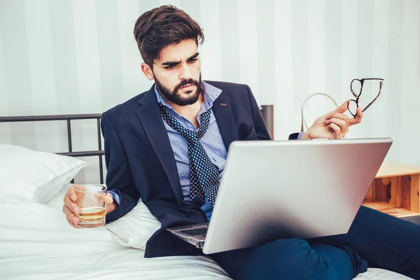 Working in hotel room. Confident young businessman in suit and tie working on laptop while sitting on the bed in hotel room