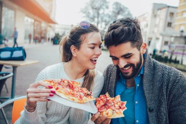 Dışarıda pizza yiyen ve gülümseyen bir çift. Açık bir kafede pizza yiyorlar..