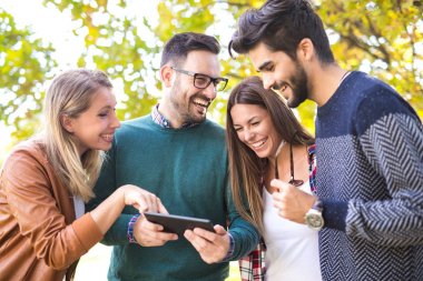 Dört genç arkadaşlar açık havada dijital tablet kullanarak Park'ta yürüyordunuz gülümseyen mutlu