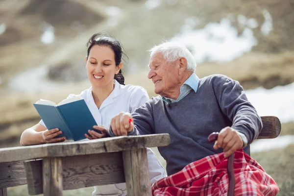 Leende Sjuksköterska Läsa Bok Till Senior Man Utomhus — Stockfoto