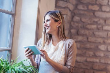 O pencereden dışarı bakarak dijital tablet aygıtı kullanarak genç gülümseyen kadın portresi