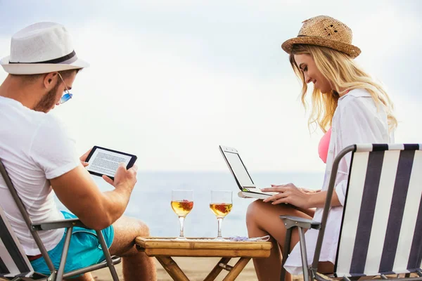 Happy smiling couple surfing net and enjoying summer at tropical beach using laptop and digital tablet