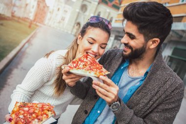 Dışarıda pizza yiyen ve gülümseyen bir çift. Açık bir kafede pizza yiyorlar..