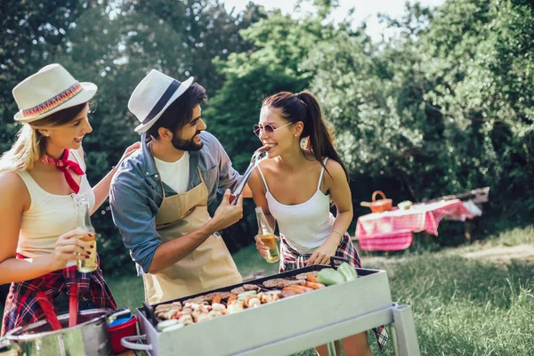 Group of friends stand at a barbecue, one cooking at grill — Stock Photo, Image