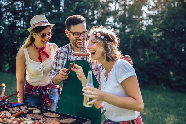 Group of friends stand at a barbecue, one cooking at grill — Stock Photo, Image