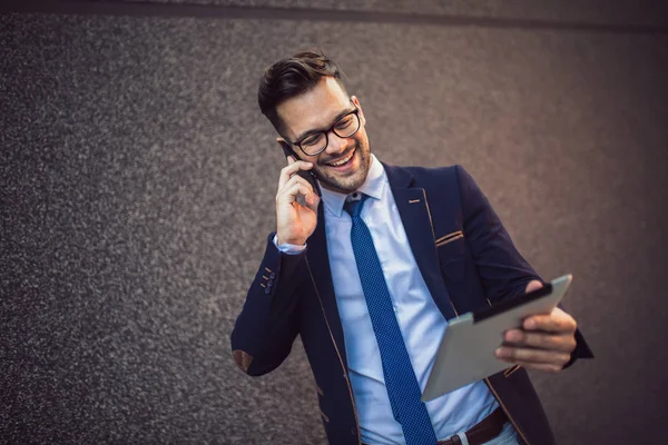 Homem de negócios sorrindo vestindo terno falando com telefone segurando tabl — Fotografia de Stock