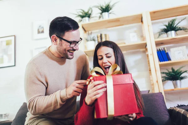 Hombre sorprendiendo a su novia con un regalo en el sofá en casa i — Foto de Stock