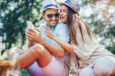 Parktaki piknik battaniyesinde içi meyve dolu piknik sepetiyle yatan çift akıllı telefon kullanıyor.