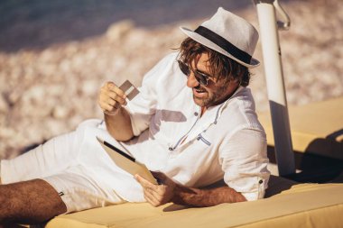 Adam deniz sahilinde rahatlıyor ve tablet bilgisayar ve kredi kartı kullanıyor.