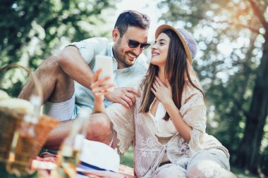 Parktaki piknik battaniyesinde içi meyve dolu piknik sepetiyle yatan çift akıllı telefon kullanıyor.