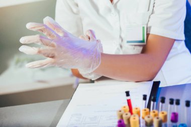 Beyaz üniformalı beyaz kadın laboratuvar asistanı laboratuarda oturuyor ve steril lastik eldivenler takıyor..