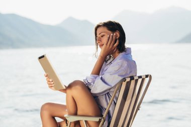 Güneşli bir günde tablet kullanarak deniz tarafından şezlongda oturan gülümseyen kadın