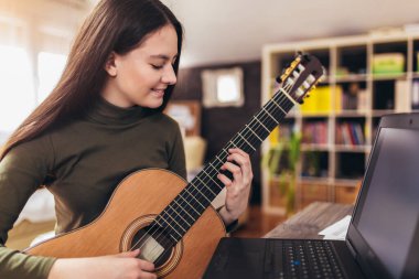 Akustik gitar çalan ve evde pratik yaparken dizüstü bilgisayarda ders izleyen bir kız. Çevrimiçi eğitim, online dersler.