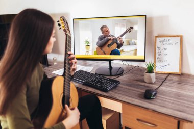 Akustik gitar çalan ve evde pratik yaparken dizüstü bilgisayarda ders izleyen bir kız. Çevrimiçi eğitim, online dersler.