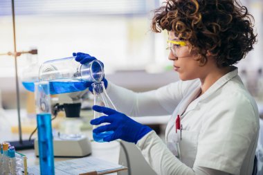 Koruyucu gözlüklü ve eldivenli genç çekici kadın bilim adamı bilimsel kimya laboratuvarında mavi sıvı madde sondasıyla test tüpü kullanıyor.