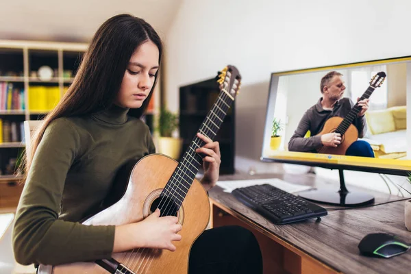 Focused girl playing acoustic guitar and watching online course on laptop while practicing at home. Online training, online classes.