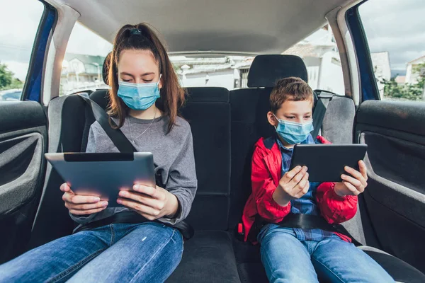 Kids wearing anti virus masks and using digital tablets in the car. Kids are travelling in car during coronavirus outbreak