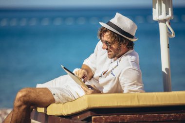 Adam deniz sahilinde rahatlıyor ve dijital tablet kullanıyor.