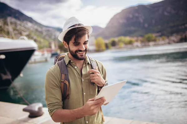 Young man walking by the harbor of a touristic sea resort with boats on background, holding digital tablet.