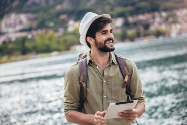Elinde dijital tabletle arka planda teknelerle turistik bir tatil köyünün limanında yürüyen genç adam..