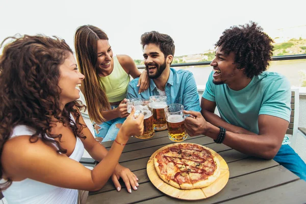 Friends enjoying pizza. Group of young cheerful people eating pizza and drinking beer while sitting at the bean bags on the roof of the building