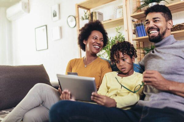 Cute little Afro-American girl and her beautiful young parents using digital tablet and doing shopping online while sitting on a sofa at home.