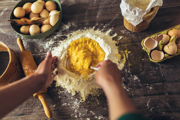 Hands whipping eggs into raw tough on wooden table.Ingredients for italian homemade pasta
