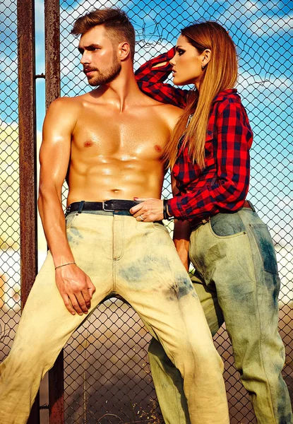 Sexy elegante loira jovem modelo com maquiagem brilhante com pele bronzeada perfeita e homem musculado bonito em jeans ao ar livre — Fotografia de Stock