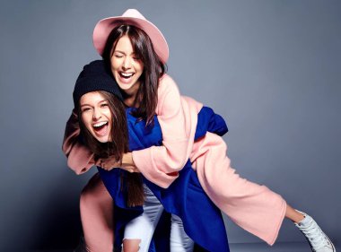 iki esmer bayan modelleri yaz casual hipster palto gri arka plan üzerinde poz içinde gülümseyen moda portre. Birbirlerine arkasında tutarak kızlar 
