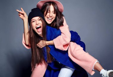 iki esmer bayan modelleri yaz casual hipster palto gri arka plan üzerinde poz içinde gülümseyen moda portre. Birbirlerine arkasında tutarak kızlar 