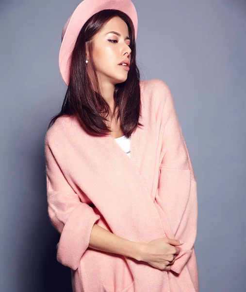 Красивая улыбающаяся брюнетка-хипстер модель в стильном розовом пальто и красочной шляпе позирует на сером фоне — стоковое фото