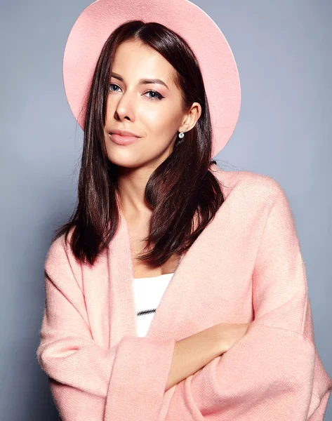 Hermoso modelo de mujer morena hipster sonriente en abrigo rosa elegante y sombrero colorido posando sobre fondo gris —  Fotos de Stock