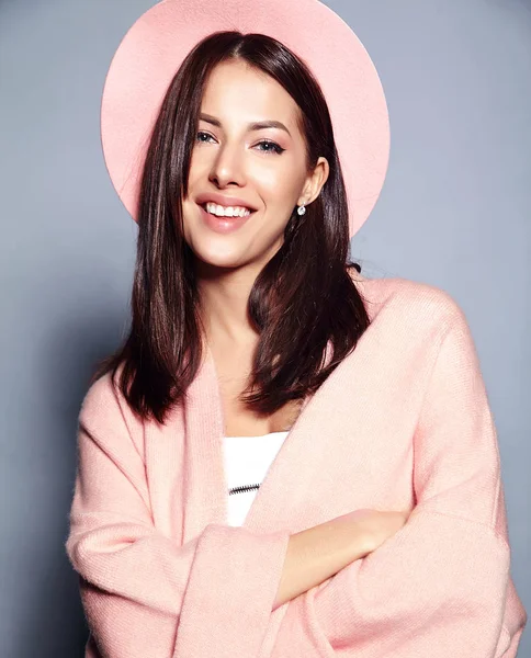 Bonito modelo de mulher morena hipster sorridente em casaco rosa elegante e chapéu colorido posando em fundo cinza — Fotografia de Stock