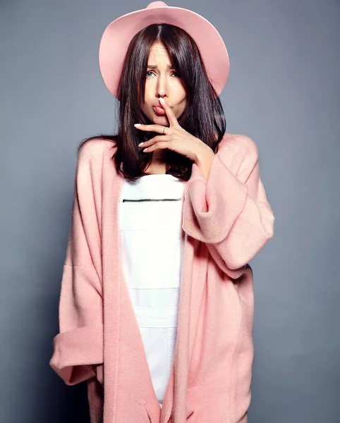 Красивая улыбающаяся брюнетка-хипстер модель в стильном розовом пальто и красочной шляпе позирует на сером фоне — стоковое фото