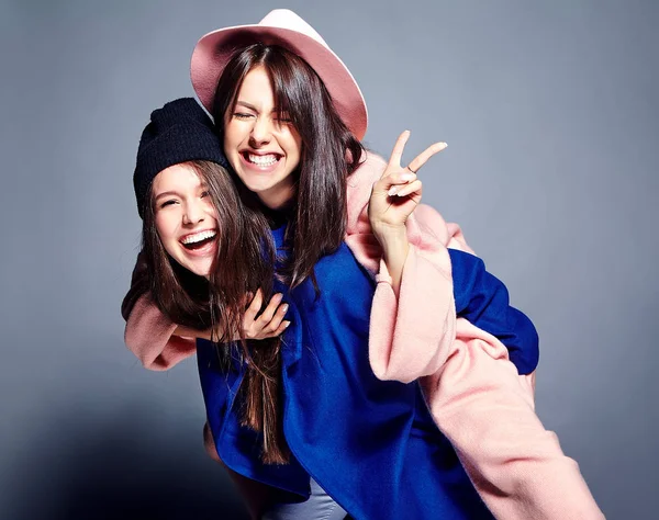 Retrato de moda de dos mujeres morenas sonrientes modelos en verano abrigo hipster casual posando sobre fondo gris. Chicas abrazándose en la espalda —  Fotos de Stock