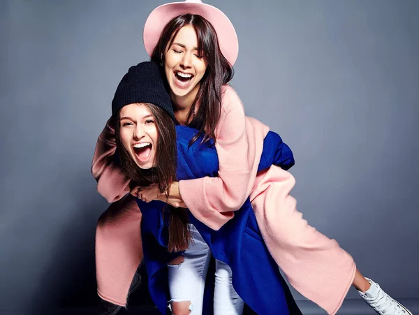 Modeporträt von zwei lächelnden brünetten Models in sommerlichem lässigem Hipster-Mantel, die auf grauem Hintergrund posieren. Mädchen halten sich gegenseitig auf dem Rücken — Stockfoto