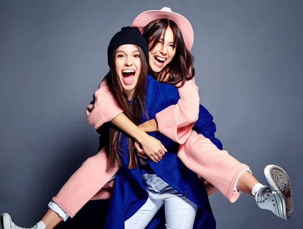 Retrato de moda de dos mujeres morenas sonrientes modelos en verano abrigo hipster casual posando sobre fondo gris. Chicas abrazándose en la espalda —  Fotos de Stock