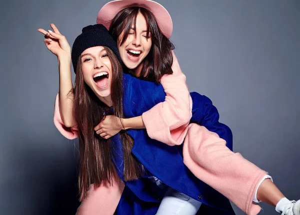 Modeporträt von zwei lächelnden brünetten Models in sommerlichem lässigem Hipster-Mantel, die auf grauem Hintergrund posieren. Mädchen halten sich gegenseitig auf dem Rücken — Stockfoto