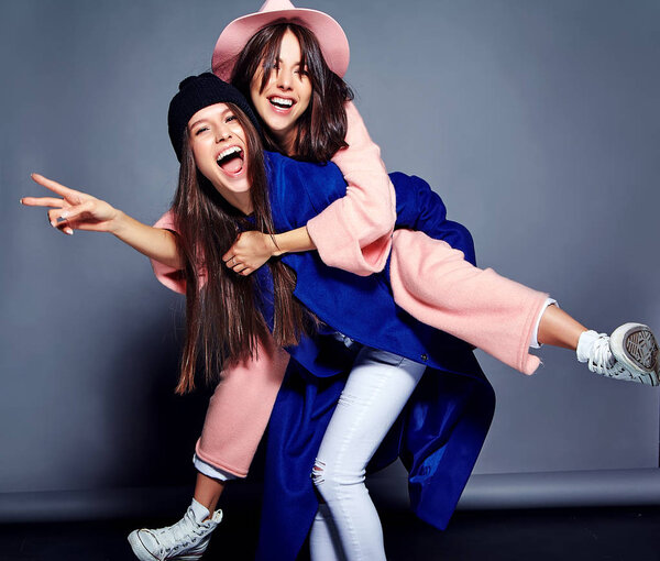 fashion portrait of two smiling brunette women models in summer casual hipster overcoat posing on gray background. Girls holding each other on back 