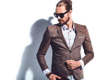 yakışıklı şık hipster işadamı manken portresi Studio beyaz duvarının yakınında poz güneş gözlüklü zarif kahverengi takım elbise giymiş. Çıkarır veya onun deri cüzdan ceket cebine attı