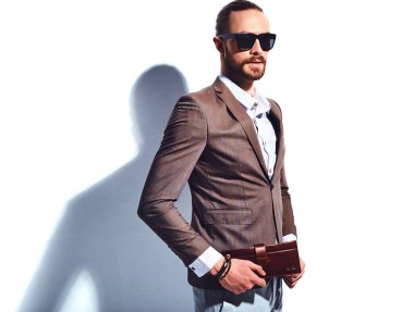 yakışıklı şık hipster işadamı manken portresi Studio beyaz duvarının yakınında poz güneş gözlüklü zarif kahverengi takım elbise giymiş. Çıkarır veya onun deri cüzdan ceket cebine attı