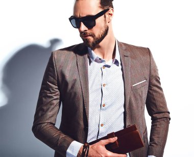 yakışıklı şık hipster işadamı manken portresi Studio beyaz duvarının yakınında poz güneş gözlüklü zarif kahverengi takım elbise giymiş. Çıkarır veya onun deri cüzdan ceket cebine attı