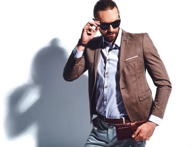 yakışıklı şık hipster işadamı manken portresi Studio beyaz duvarının yakınında poz güneş gözlüklü zarif kahverengi takım elbise giymiş. Çıkarır veya onun deri cüzdan ceket cebine attı