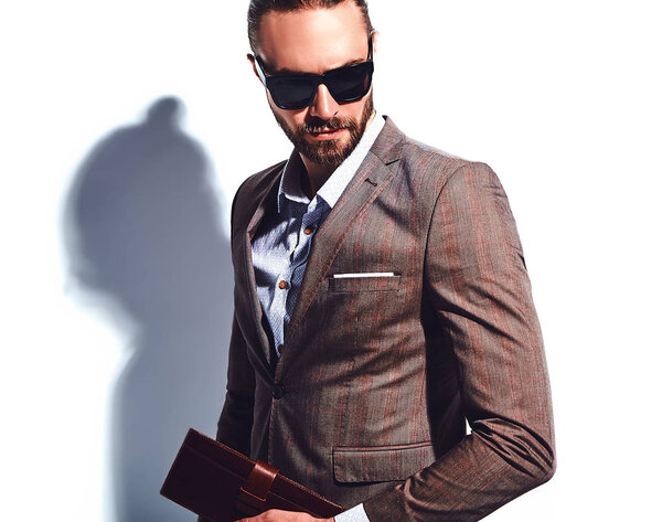 portrait of handsome fashion stylish hipster businessman model dressed in elegant brown suit in sunglasses posing near white wall in studio. Pulls out or put his leather wallet into jacket pocket